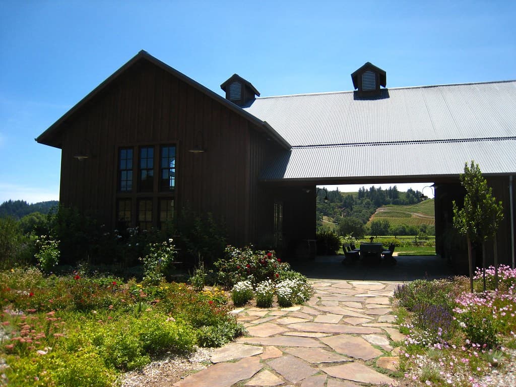 Dark wood barn tasting room and breezeway behind a flowering garden and flagstone path at Dutcher Crossing
