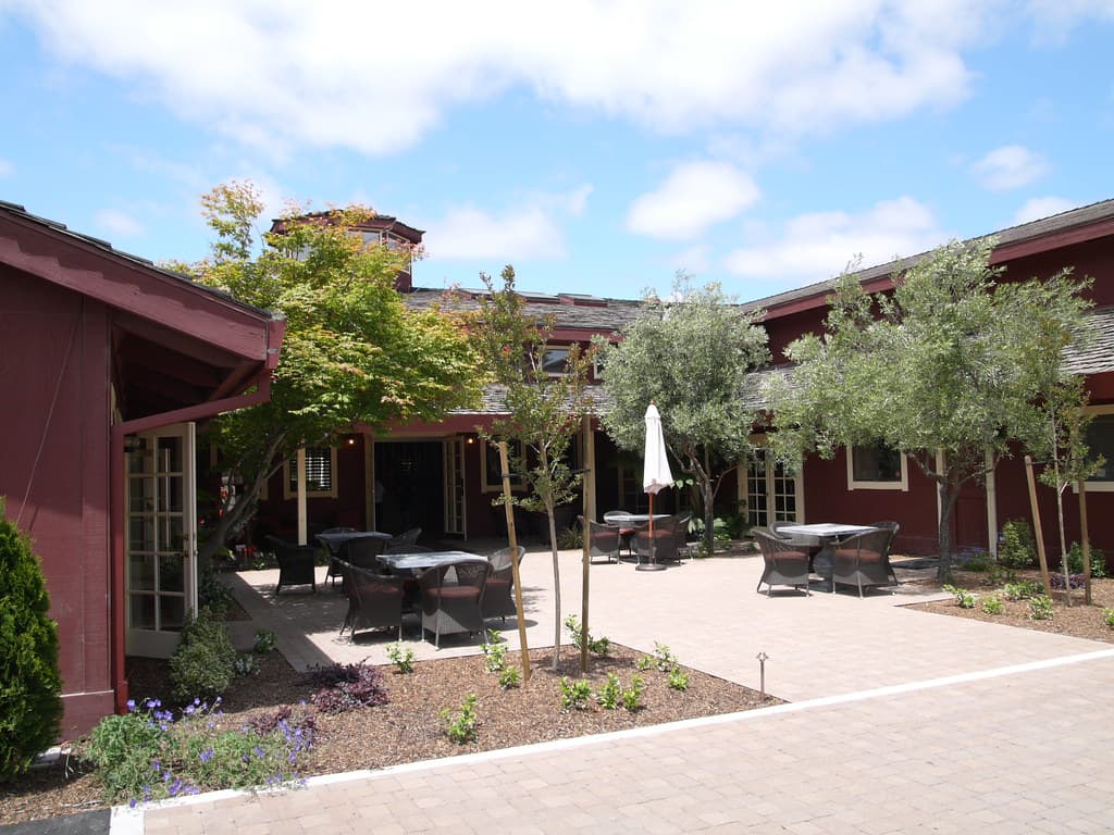 Brick courtyard with patio tables, umbrellas and young olive trees between the red barn buildings at DeLoach Vineyards