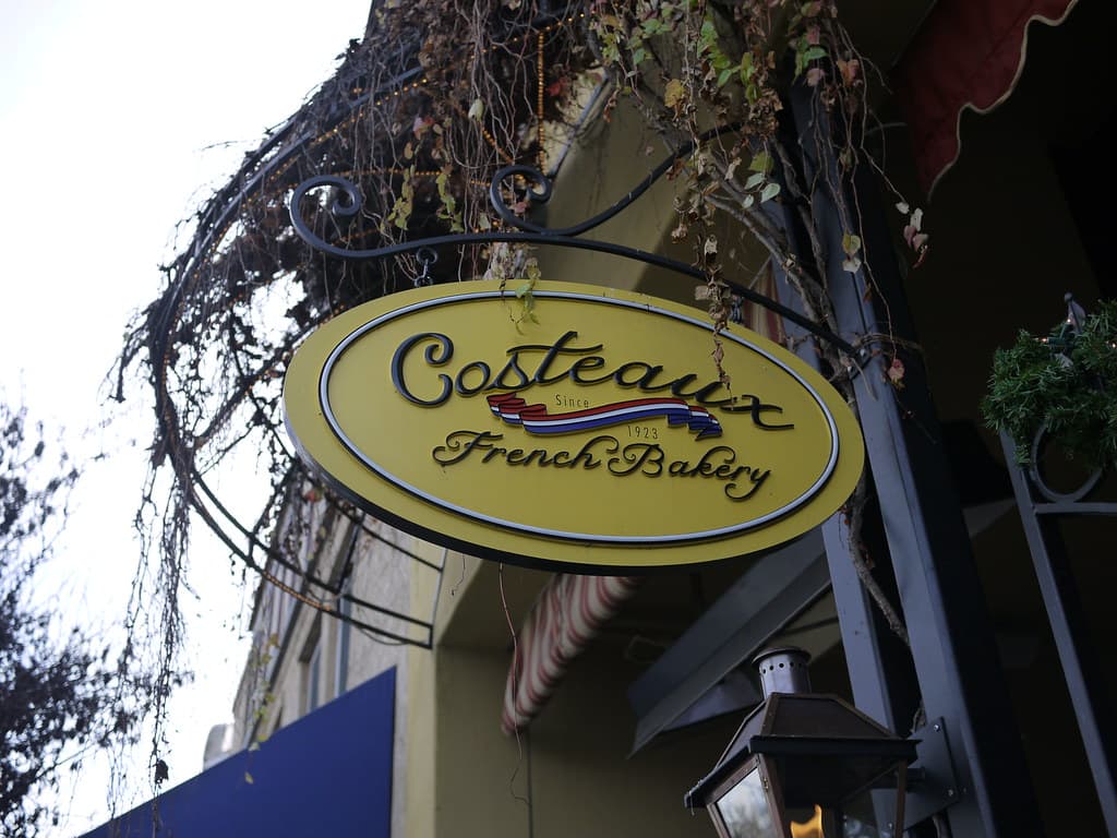 The yellow oval Costeaux French Bakery sign hanging from an iron bracket over the bakery's entrance