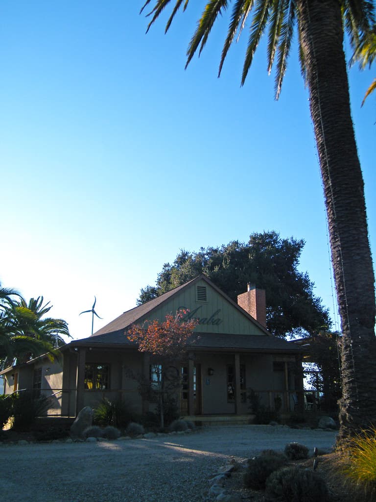 Farmhouse-style Anaba tasting room under oaks and a palm, with a small wind turbine behind