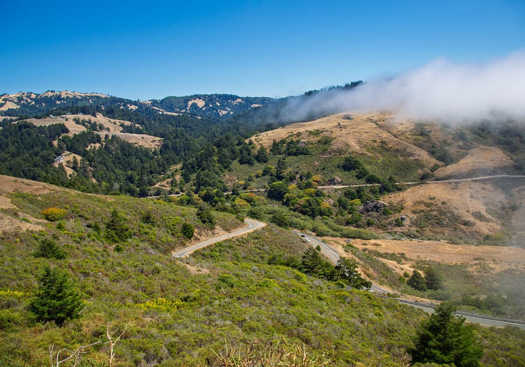 Highway 1 winding down through scrub-covered hills into Russian Gulch north of Jenner, with fog drifting over the ridges