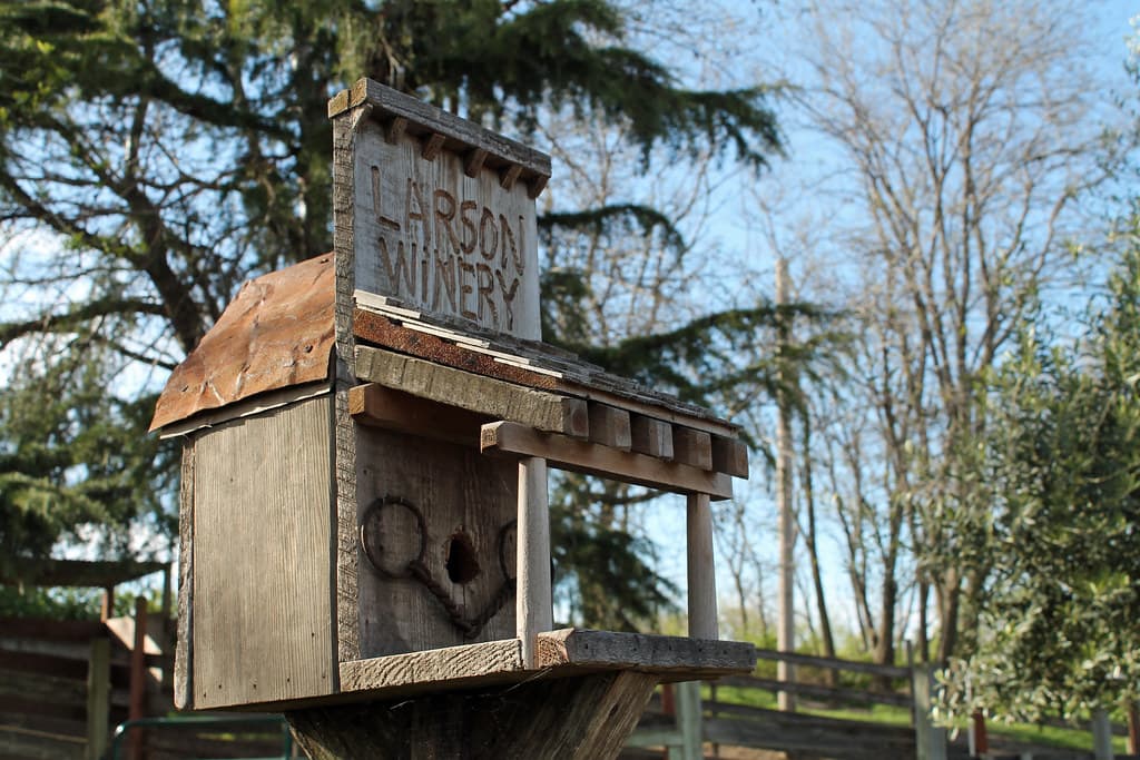 Photo: sarahstierch (CC0) Rustic hand-built Larson Winery sign with a weathered roof and horseshoe, under oaks at the ranch entrance
