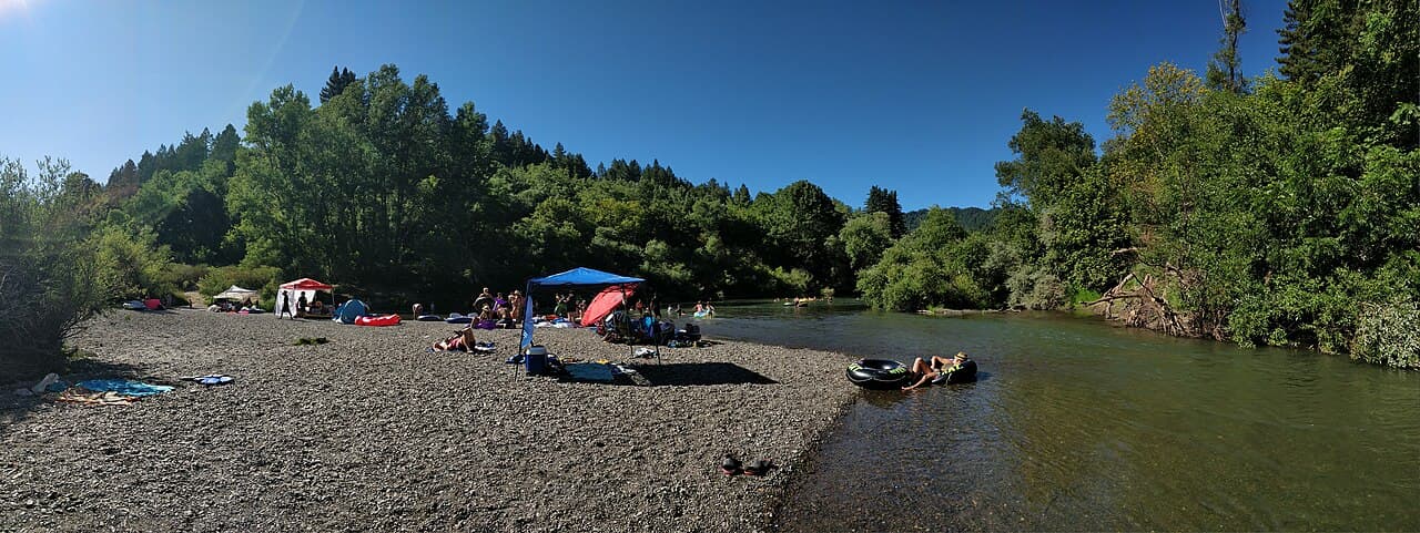 Panorama of the gravel river beach at Sunset Beach with sunshades, swimmers and wooded banks along the Russian River