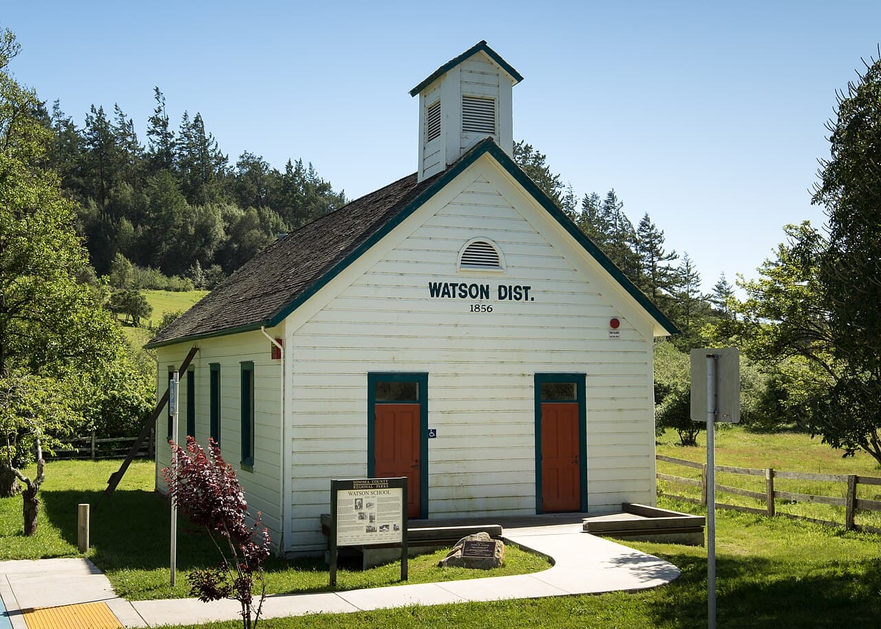 The white 1856 Watson District one-room schoolhouse with its cupola, lawn and interpretive sign at Running Fence Watson School Historic Park