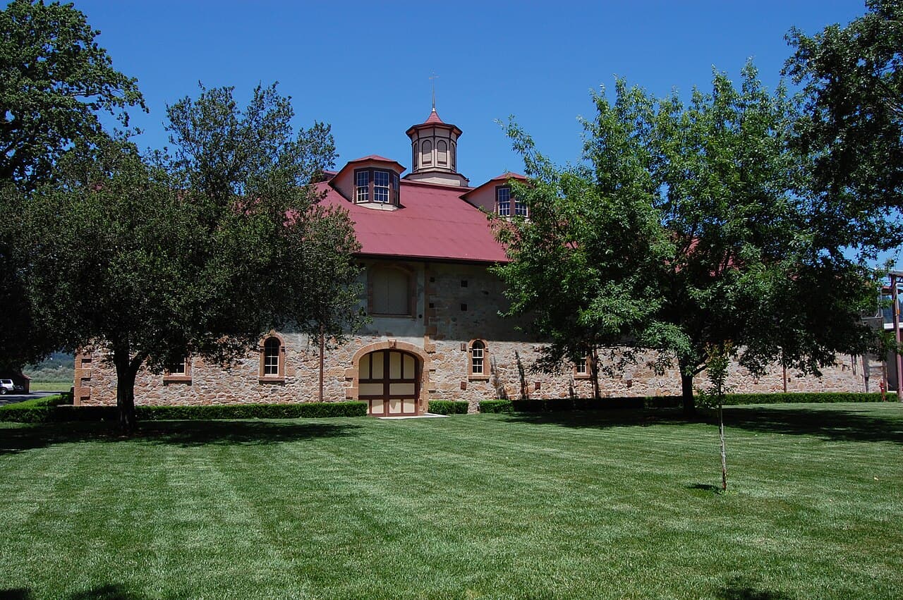 Striped green lawn and shade trees in front of the historic stone winery with its red roof and cupola at Charles Krug