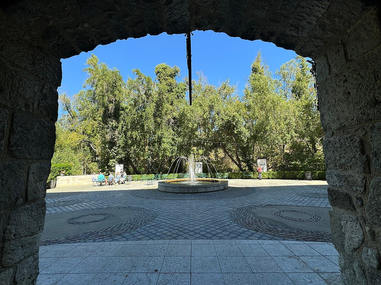 Cobblestone courtyard with a round fountain and bistro tables under tall trees, seen through a stone archway at Buena Vista Winery
