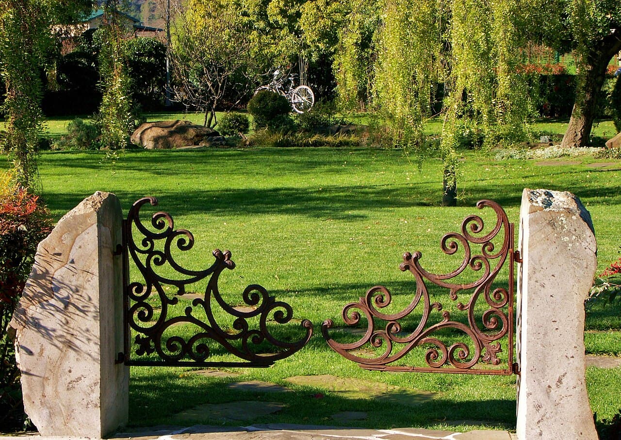 Ornate iron gate between rough stone pillars opening onto a sunny lawn and willow-shaded garden at Peju Winery