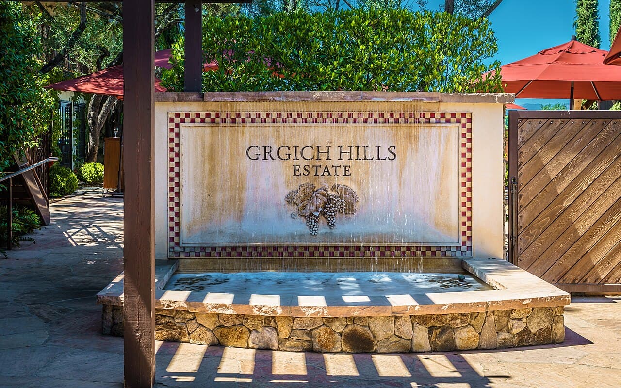 The tiled Grgich Hills Estate sign above a stone fountain, with red umbrellas and greenery behind it in Rutherford