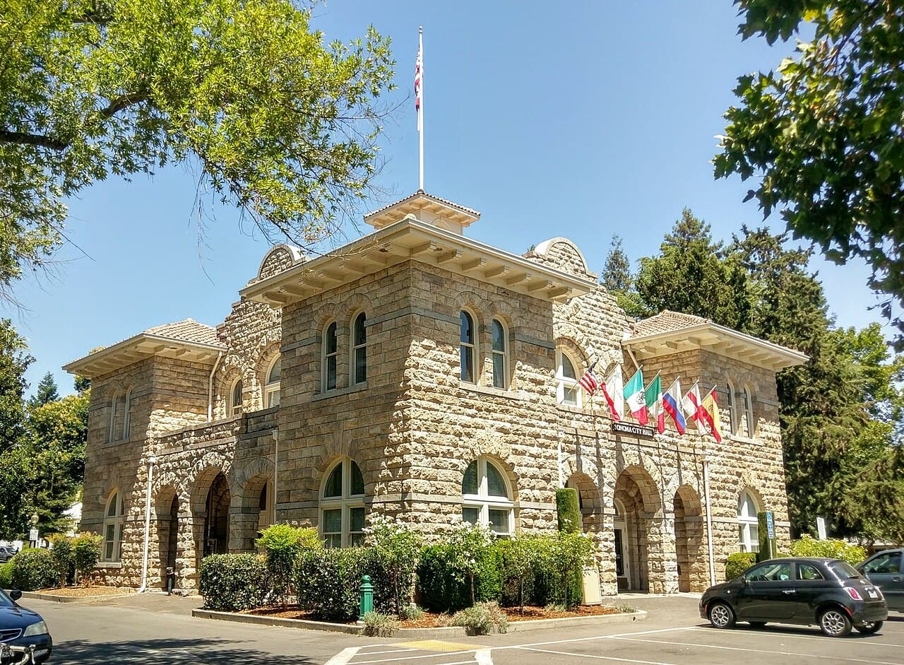 Sandstone Sonoma City Hall with flags, framed by trees at the center of Sonoma Plaza, where the Tuesday Night Market is held