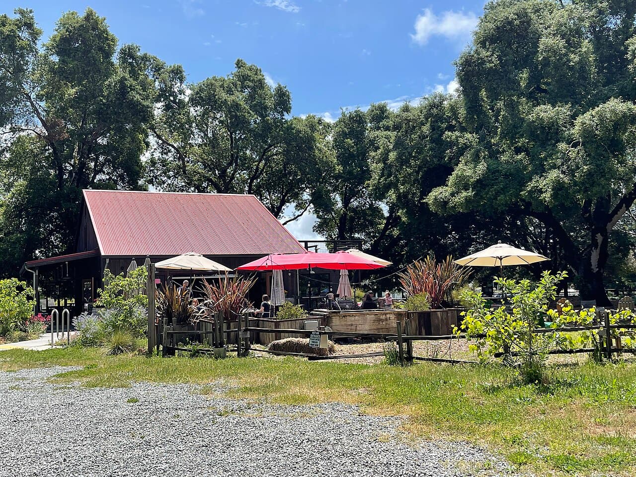 Red and cream umbrellas shading outdoor tables beside the red-roofed redwood barn under big oaks at Horse & Plow