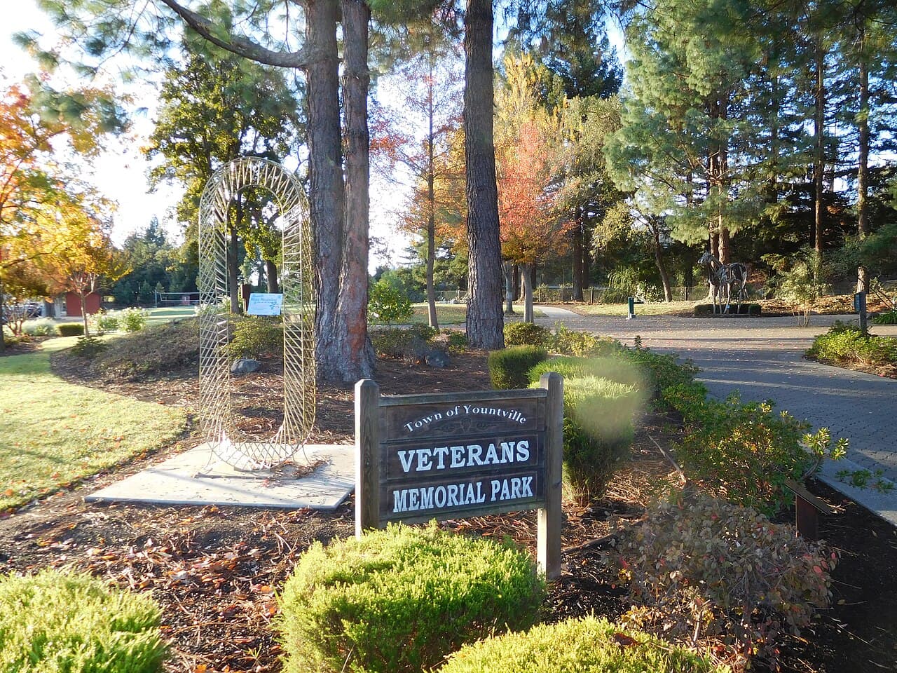 Town of Yountville Veterans Memorial Park sign beside a wire arch sculpture, tall pines, autumn trees and a brick path