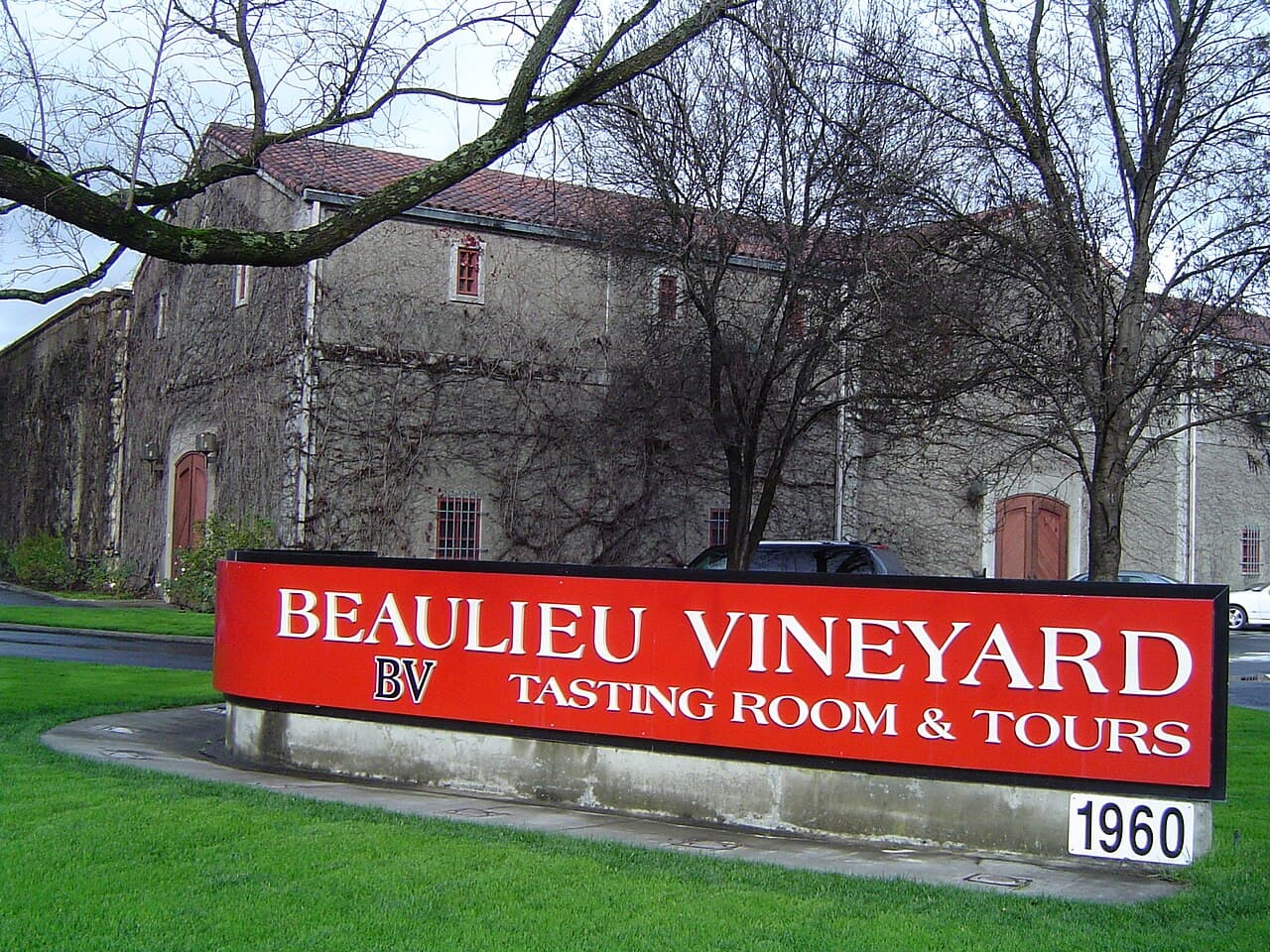 Red Beaulieu Vineyard BV tasting room sign on a lawn in front of the ivy-bare historic stone winery building under winter trees