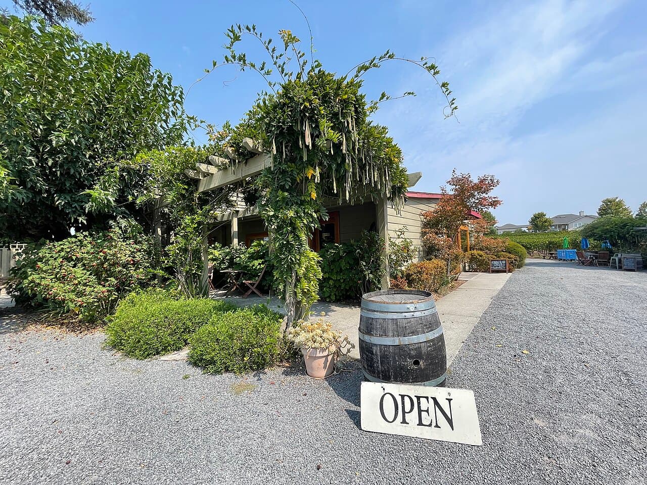 Wisteria-covered pergola and garden at the Furthermore Wines tasting room, with a barrel and open sign on the gravel drive